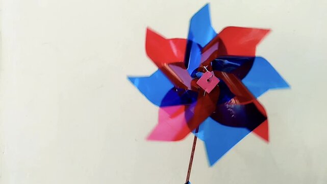 Footage of a glossy plastic pinwheel in red and blue spinning naturally in the wind with a cream painted wall background. ideal for childhood, education, renewable energy concepts, outdoor lifestyle
