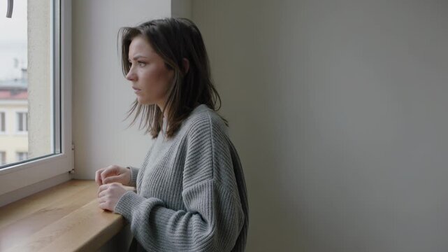 Leaning adult woman in grey sweater scanning outside seeing motion at window, reaching for latch