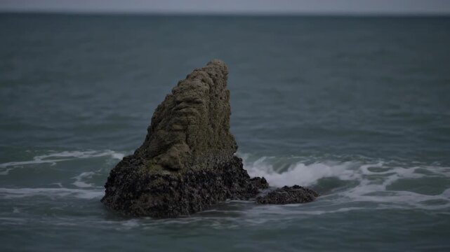 Swell arriving at sea, rock outcrop with barnacled base enduring waves, revealing nearby reef