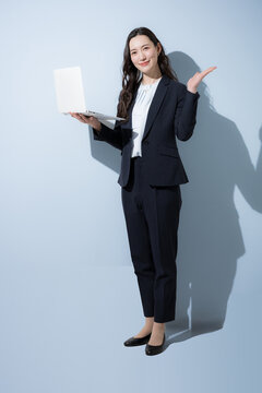 A Japanese businesswoman in a suit, smiling as she types on a laptop against a light blue background; a full-body business portrait evoking images of DX, AI, operational improvements, and IT services