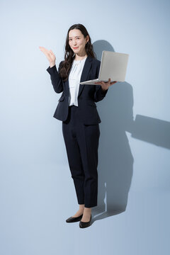 水色背景でノートパソコンを持つ笑顔でタイピングするスーツの日本人ビジネス女性、DXやAI、業務改善、ITサービスをイメージしたビジネスポートレート全身　カメラ目線のオススメや紹介のポーズ