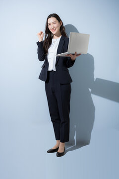 水色背景でノートパソコンを持つ笑顔でタイピングするスーツの日本人ビジネス女性、DXやAI、業務改善、ITサービスをイメージしたビジネスポートレート全身　カメラ目線のガッツポーズの元気や成功ポーズ