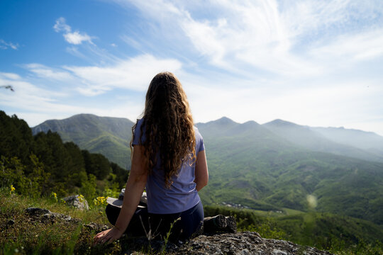 Rear view of a female traveler sitting quietly on a mountain viewpoint surrounded by scenic summer nature.