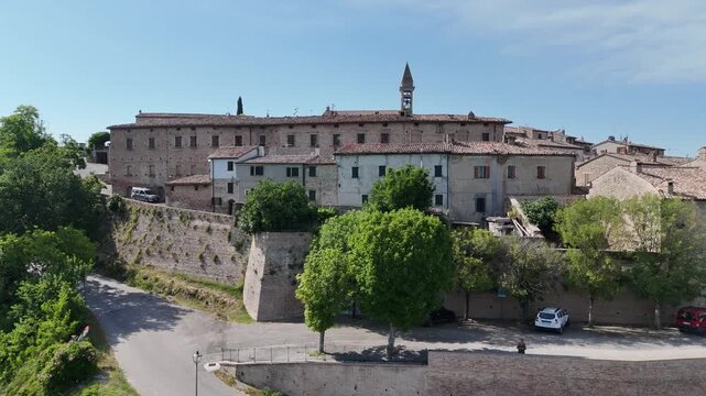 Borgo storico in cima a una collina con edifici in pietra, campanile dominante e vegetazione circostante, sotto cielo limpido. Atmosfera tradizionale e mediterranea