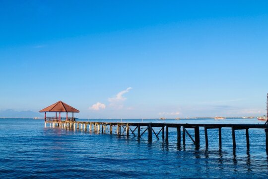 Wooden pier with gazebo over calm blue sea at Tanjung Lesung Beach, Banten, Indonesia. Wide-angle coastal view under clear sky, creating a peaceful escape. Ideal for travel, tourism, summer, and leisu