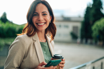 Young businesswoman smiling, using phone, drinking coffee in a city environment, enjoying outdoor...
