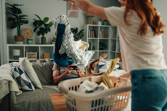 Woman throwing laundry at man ignoring chores, playing on phone, showing relationship frustration and unequal housework