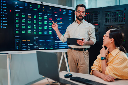 It professional explaining server rack alert status on a dashboard to a female colleague in a data center