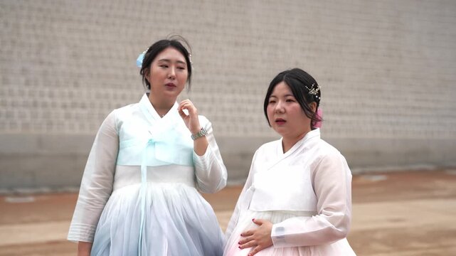 Two women in traditional Korean attire stand side by side. They appear amused, reacting to something unseen. Their gestures and expressions convey lighthearted joy and playful surprise