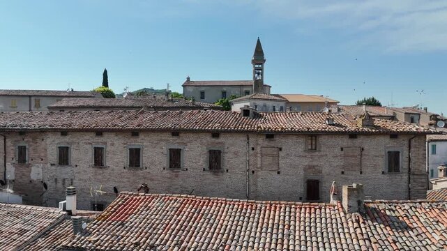 Villaggio rurale con edifici in pietra e tetti in coppi, dominato da un alto campanile, circondato da colline verdi e campi coltivati sotto cielo azzurro limpido