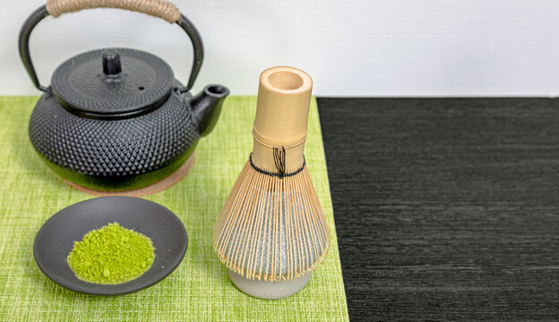 Matcha tea powder in small bowl with bamboo whisk and black iron kettle. Traditional Japanese tea ceremony equipment on green mat. Healthy beverage preparation and organic food concept