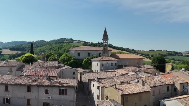 Villaggio rurale con edifici in pietra e tetti in coppi, dominato da un alto campanile, circondato da colline verdi e campi coltivati sotto cielo azzurro limpido