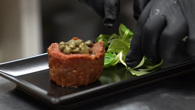Skilled chef wearing black gloves carefully placing fresh corn salad next to beef tartare