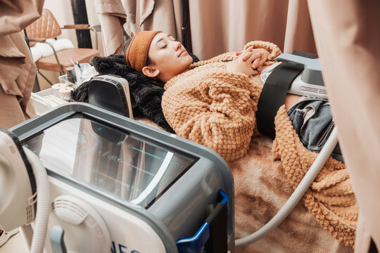 Young Asian woman relaxing during a High-Intensity Focused Electromagnetic or HIFEM body contouring treatment targeting the abdominal muscles at a modern aesthetic clinic.