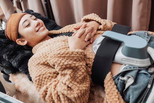 Young Asian woman relaxing during a High-Intensity Focused Electromagnetic or HIFEM body contouring treatment targeting the abdominal muscles at a modern aesthetic clinic.