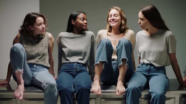 Turning second-left woman initiating chat, four women shifting on metal counter in studio, jeans