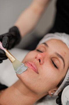 Beauty therapist applying a grey facial mask to a client lying on a treatment bed