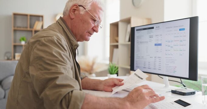 Senior elderly man checks home electricity bill. At a computer with utilities invoice, he prepares payment and reviews expenses to manage a balanced budget. Clear concept of household savings.
