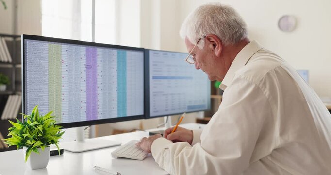 Senior accountant man analyst at office computer. He reviews spreadsheet finance data on dual monitors, noting and confirming analysis for reporting. Concept of precise financial analytics.