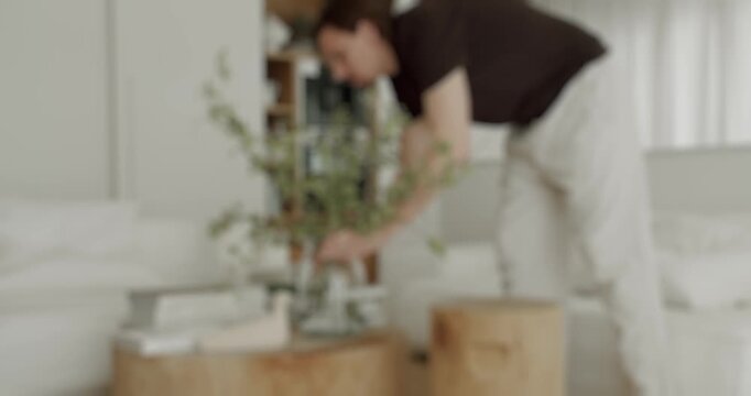 Blurry soft focus video of a young man arranging fresh green foliage in a glass vase on a wooden coffee table, bringing nature and minimalist style into his bright and modern home interior