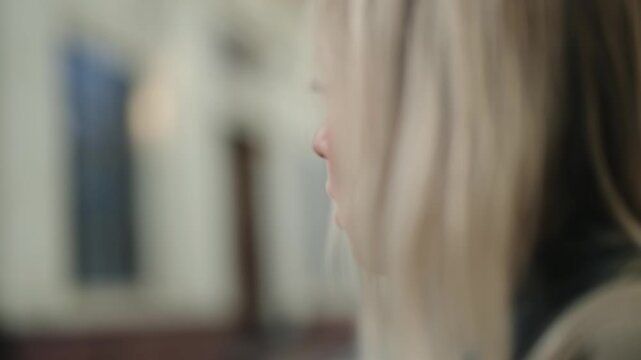 Profile view of a young blonde woman vaping with an electronic cigarette, holding the device in her hand and inhaling the vapor in a room with a shallow depth of field, filmed in slow motion