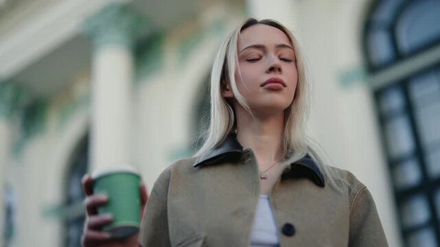 Slow motion low angle portrait of a stylish confident young blonde woman holding a takeaway coffee cup while walking down a beautiful city street and looking around with a serious expression