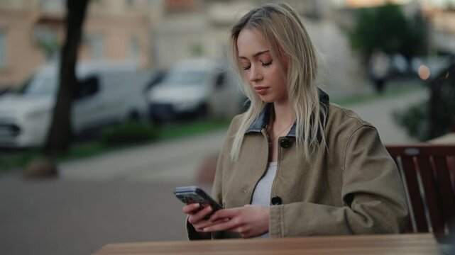 Beautiful young blonde woman sitting alone at a table in a city cafe, focused on her smartphone screen while scrolling through social media or texting with friends on a cloudy day