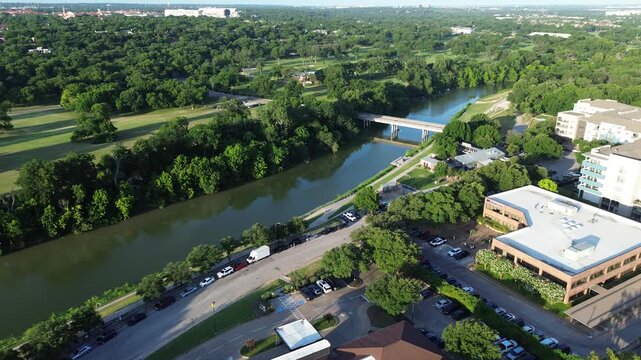 Lush tree lined banks border Trinity River, flowing beneath bridge near office suites. Vehicles navigate streets between mature vegetation, tree canopies, parking lot perimeters, Fort Worth, Texas