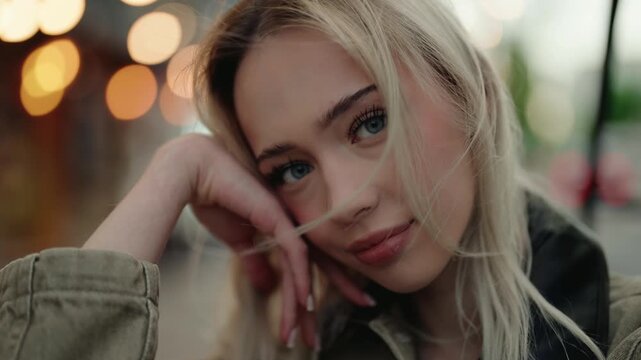 Charming young blonde woman with beautiful blue eyes looking at the camera with a gentle smile on a city street at night, her hair blowing in the wind in slow motion with bokeh lights