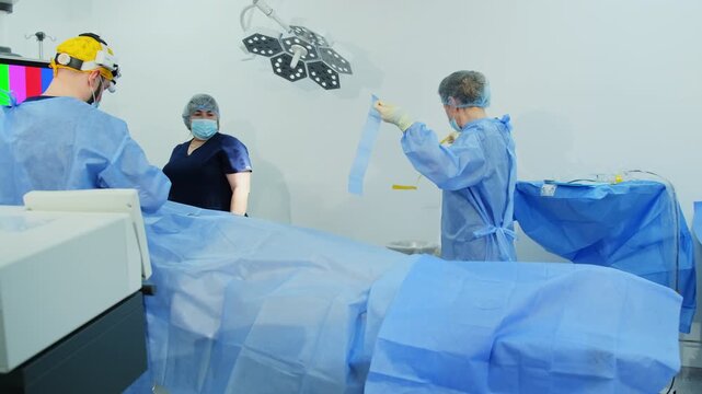 Diligent operating nurse carefully secures sterile surgical drape on patient. Women prepare bright operating room for surgery. Slow motion