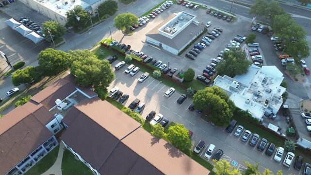 Asphalt lots define retail, professional hub near major road intersections, leafy neighborhoods. Mature trees cast morning shadows over vehicle rows, low rise building structures, Fort Worth, TX