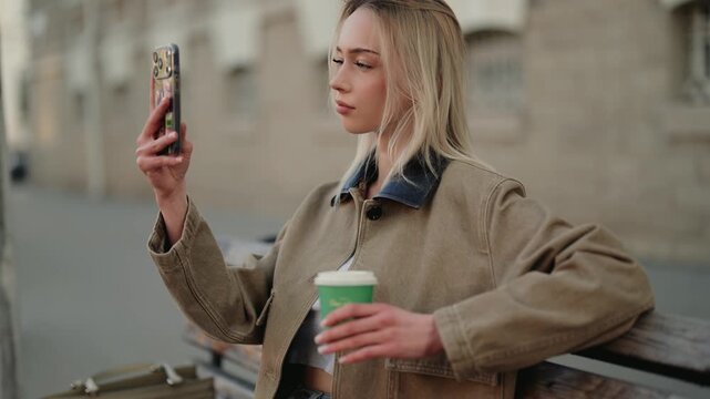 Attractive young woman sitting on a city bench, taking a selfie with her smartphone while holding a cup of takeaway coffee, enjoying a relaxing moment during her break outdoors in slow motion