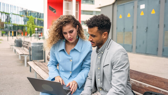 Two business professionals using a laptop on a city bench, collaborating on a project or discussing work in an urban setting