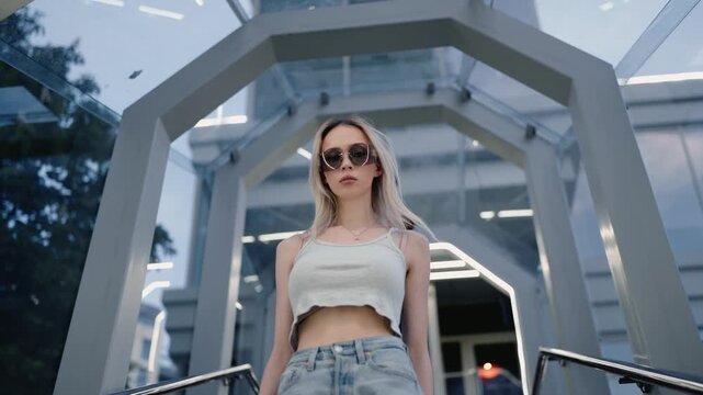 Confident young woman with blonde hair wearing sunglasses walking up the stairs from a modern subway station exit, looking directly at the camera in a contemporary urban environment