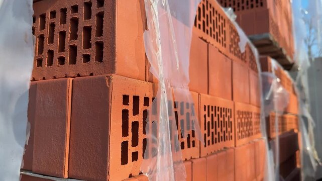 Close-up stacked red bricks hardware store, street side pallet of hollow clay blocks wrapped in plastic, sunlight highlighting rough