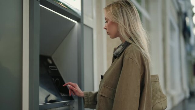 Young blonde woman using a credit card and touchscreen to withdraw money from a modern ATM machine located on a city street, completing a secure financial transaction