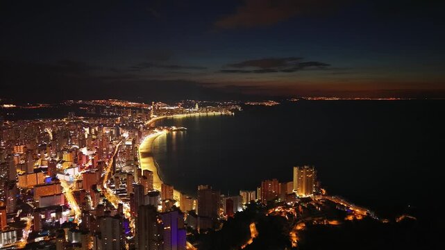 Twilight aerial coastline bathing in afterglow and soft city lights, wide panorama of crescent bay, gentle