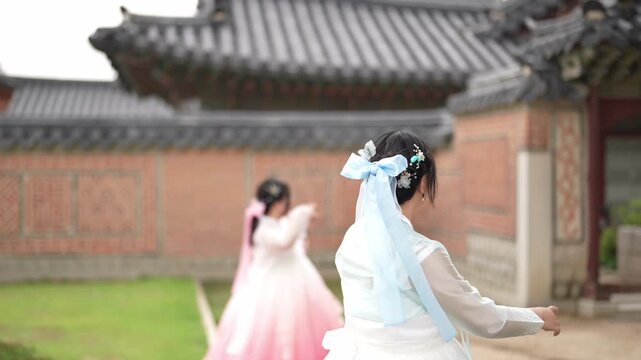 Young girls in traditional Korean attire play joyfully outdoors. They laugh, dance, and move freely amid historic architecture. Bright colors and soft focus emphasize their carefree, happy expressions