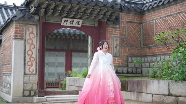 Woman in a pink hanbok walks past a traditional Korean gate. Her smile suggests calm joy, perhaps during a cultural visit. The gate features intricate tile patterns and stone architecture