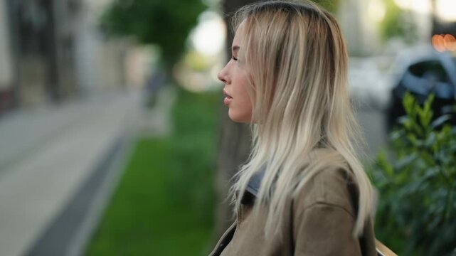 Profile view of a young woman with long blonde hair smoking an electronic cigarette and exhaling vapor while sitting on a bench outdoors in a city park