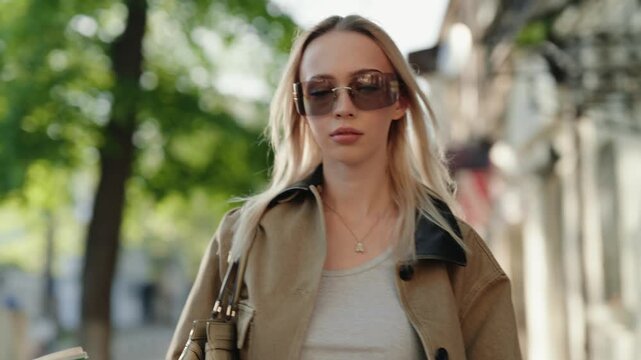 Confident young blonde woman in sunglasses walking down a sunny city street holding a cup of coffee, her hair blowing in the wind while she looks around at the urban scenery in slow motion