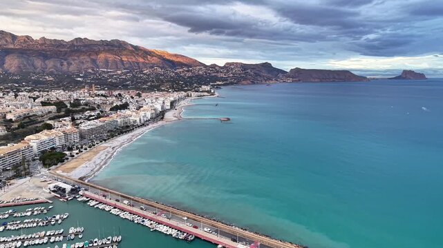 Aerial marina town with turquoise water and pier, calm surf along sandy beach, mountains and clouds shaping