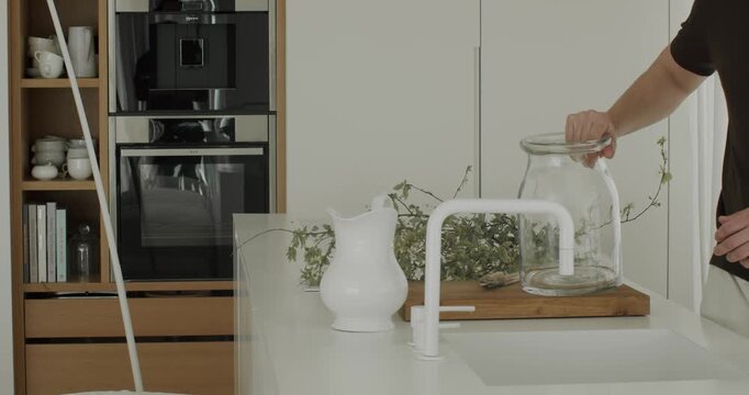 Unrecognizable man removing green branches from a large glass vase and filling it with fresh water from a modern white faucet in a stylish, contemporary kitchen with built-in appliances