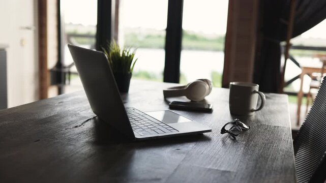 A laptop, headphones, smartphone, eyeglasses, and a mug are arranged on a wooden table near a window