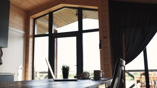 A laptop, headphones, and coffee mug sit on a wooden table in a rustic cabin with large windows offering a bright outdoor view