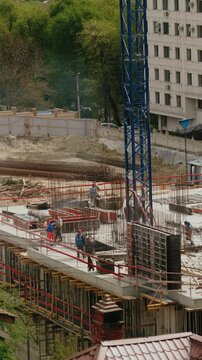 Team of builders working on a reinforced concrete structure at a construction site, with a large blue crane nearby and lush green trees and an office building in the background. timelapse. vertical