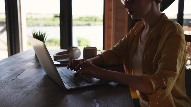 A caucasian woman wearing glasses and a yellow shirt works on her laptop at a wooden table with a scenic view of a lake and greenery outside