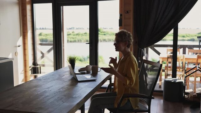 A caucasian woman with short blonde hair and glasses is engaged in a video conference call while sitting at a wooden table in a room with large windows overlooking a serene lake