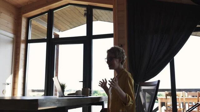 A woman with short curly hair and glasses waves hello while participating in a video conference call on her laptop in a wooden cabin