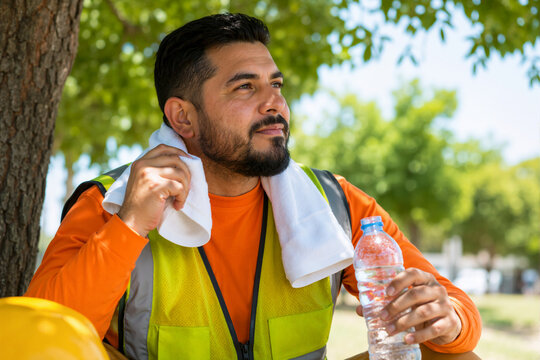 Construction worker resting in the shade and wiping sweat with a towel. Hispanic man in reflective vest holding a water bottle to prevent heat stroke. Occupational safety and hydration concept